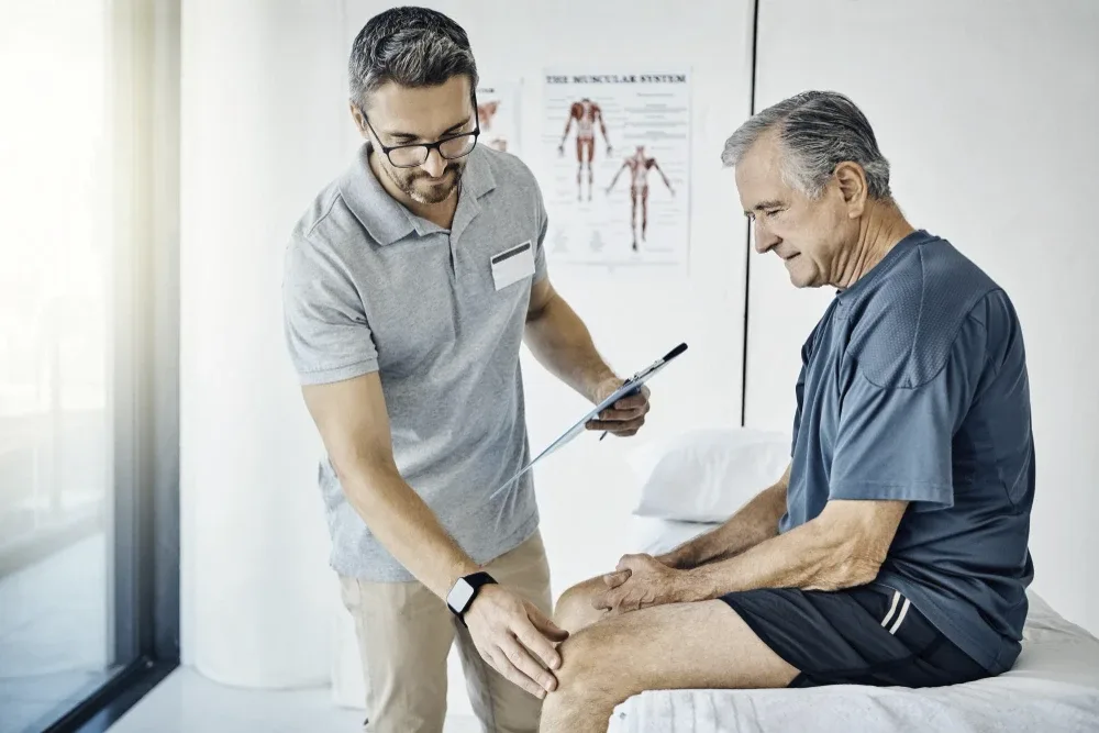 A clinician examining an older man's knee in a consulting room