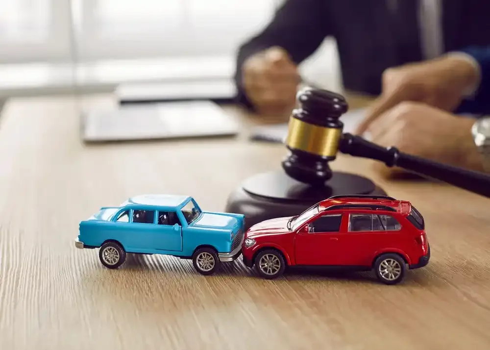 Dwa zabawkowe samochodziki ustawione tak, jakby doszło do zderzenia tylnego, na biurku obok młotka sędziowskiego